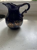 Dark blue ceramic creamer with pheasant and floral gold design, decorative handle and spout