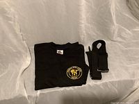 Black Poseidon T-shirt folded with shoulder strap next to it, showing logo on chest.