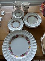 Photo of dinner set with stacked plates in three sizes and four cups with floral pattern and ruffled edges, showing condition and design details.