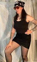 Female model wearing black tank, black suede mini skirt, fishnet pantyhose, tattoo print nylon sleeves, black belt, black and white do rag, and sunglasses.