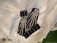 Pair of black skeleton gloves with white skeleton bone design, medium size, laid flat on white fabric. Two black plastic wristbands with large grey spikes and silver studs, with velcro closures, shown with one on wrist to depict size.