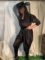 Model wearing the black satin-like jumpsuit with deep plunging neckline, 3/4 length puffed sleeves, and gathered legs, paired with the black witch hat with veil and the black and burgundy belt.