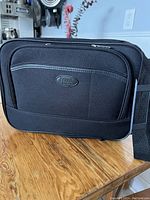 Front view of black Protocol laptop carry case on wooden surface showing the Protocol logo and front pocket.