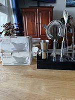 Photo showing two boxed white ceramic Maywell Williams White Basics gravy boats and a black stand holding various metal bar accessories such as a strainer, jigger, and spoons.