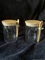 Two clear glass jam jars with wooden lids and attached wooden spoons on black cloth background