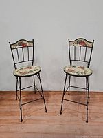 Pair of black metal folding bar stools with mosaic tile seats and backs showing floral and fruit patterns.