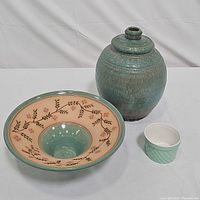Three green ceramic pottery items: lidded jar, serving platter with branch design, and seafoam ramekin arranged on white surface.