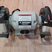 Front view of the 8 inch slow speed bench grinder showing the grinding wheels, protective shields, clear safety guards, and the power switch.