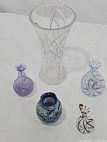 Five blown glass items placed on a white surface: a large clear etched vase, a teardrop blue swirled vase, a small purple bud vase, a round ripple vase with blue and green swirls, and a twisted pattern pitcher.