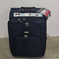 Front view of the navy blue Ricardo Elite suitcase showing the fabric, zippers, handle, and attached tags.