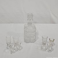Front view of crystal glass decanter with stopper and 8 assorted clear glass shot glasses arranged around it.