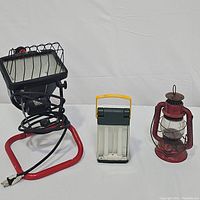 Three lighting items side by side: red kerosene lantern, Energizer folding fluorescent lantern, and halogen work light with red frame and wire grill.