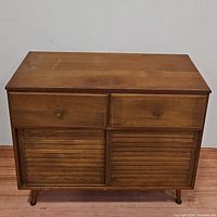Front view of mid-century modern wooden cabinet with two drawers and a sliding panel door beneath, resting on wooden legs.