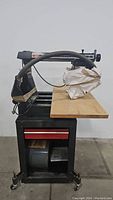 Front side view of Sears Craftsman radial arm saw on stand with dust collection hose and wood table.