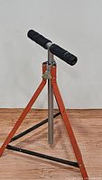 Full view of the tripod with orange metal legs and black double roller top, standing on a flat surface against a white wall.