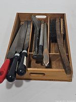 Wooden divided tray containing nine files with mostly black handles, three chisels, and two bristle brushes with wooden handles.