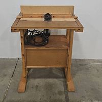 Front view of handmade wooden work table showing sturdy legs, lower shelf, and black cord coiled underneath work surface.