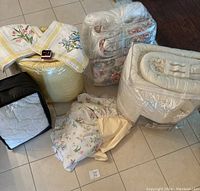 Photo shows multiple queen size bedding items including comforters, mattress pad in plastic wrap, floral bed skirt, folded bedding piece, and a jewelry box with necklace and earrings on top of a bin.