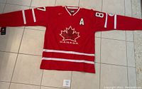 Full front view of red Canada hockey jersey laid flat showing white stripes, large signed maple leaf crest, assistant captain ‘A’ patch, and number 87 on sleeves.