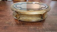 Wooden oval coffee table on hardwood floor showing the glass top and wooden lower shelf, legs visible.