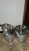 Photo showing 4 pots stacked and a metal baking tray with colander on top, various sizes and aluminum/stainless steel materials.