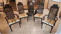 Group of six wooden dining chairs with cane backs, four without armrests, two with armrests, floral patterned cushions, and ornate carved tops.