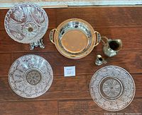 View of kitchenware items arranged on wooden floor: cut glass bowl with silver tone metal stand, brass canter, silver serving platter, and three decorative glass plates.