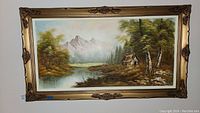 Full front view of the original signed landscape painting on canvas featuring a cabin by a river with mountains and trees, in an ornate gold frame.