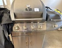 Front view of stainless steel Life Home gas grill showing control knobs, lid with thermometer, and lower cabinet storage.