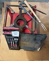 Red metal frame propane burner with circular cast burner, Snap-on BBQ utensil set box, two metal cooking trays, BBQ fork and tongs on top.