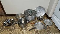 Full set of kitchen cookware on floor showing all pots, pans, bowls, colander and wire strainer with wooden handle.