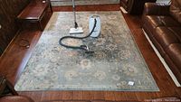 Large area rug shown laid flat on hardwood floor with vacuum cleaner placed on top, displaying pattern and scale