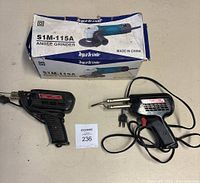 Angle Grinder S1M-115A with original box alongside two Weller soldering irons with cords and plugs, displayed on table