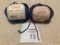 Two balls of LB Collection cashmere yarn in teal blue and purple colors with labels showing fiber content and care instructions, placed on a textured cream surface.