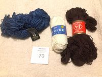 Three balls of yarn in dark blue, white, and dark brown with labels visible, placed on a light surface with a MaxSold auction card showing number 70.