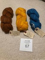 Three skeins of Quo Vadis yarn in brown, yellow, and blue laid on a light surface, each with a handwritten tag describing the yarn composition and weight.