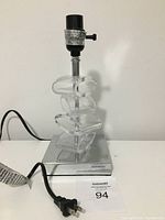 Front angle view of clear acrylic/lucite table lamp with black socket, adjustable clear pieces stacked on chrome base, and power cord visible.