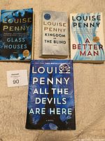 Front covers of four Louise Penny mystery novels titled Glass Houses, Kingdom of the Blind, A Better Man, and All the Devils are Here arranged on fabric surface.