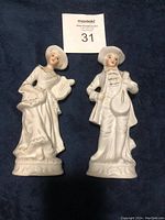 Two white porcelain Victorian style figurines, a woman with harp and a man with mandolin, standing side by side on a dark cloth background.