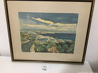 Framed watercolor painting depicting a coastal scene with lighthouse, ocean, rocks and sky.