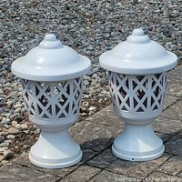 Photo showing two large white painted outdoor lanterns side by side on paved surface with a gravel area behind