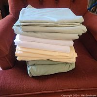 Four folded sets of queen sheets in green, yellow/gold, and white colors stacked on a chair.