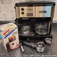 Front and angled views of DeLonghi combination coffee and espresso maker with glass carafe and espresso accessories placed in front and beside machine, next to boxed Black and Decker grinder.