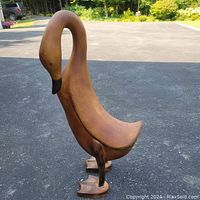 Front side view of large brown duck decorative figure showing its curved neck and black-tipped beak, standing on two webbed feet on asphalt surface.