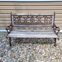 Front view of wooden and metal outdoor park bench on gravel, showing wood seat slats and ornate metal backrest and armrests.