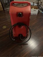 Red 14 gallon gas can with black hose coiled and attached, showing the handle and cap on a dark wood floor indoors.