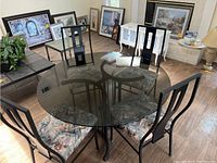 5 piece dining set including a round smoked glass table and four chairs in a room with paintings and other furniture.