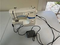Brother LS-2125 white and blue sewing machine on table, showing machine body, stitch selection dial, foot pedal, and power cord.