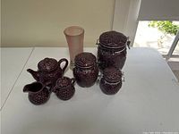 Six-piece set of dark purple ceramic kitchen canisters with lids and matching tall vase, all featuring grape cluster embossed design.