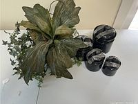 Four black and gray canisters of varying sizes with silk plant lids situated on a white surface against a beige wall.